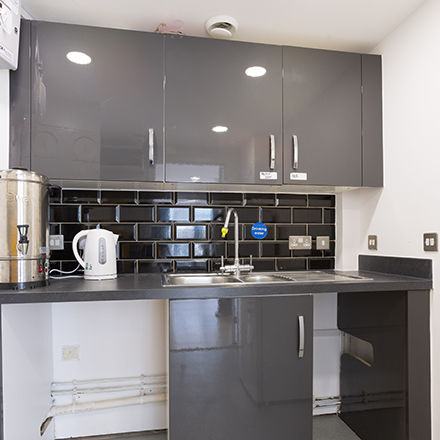 Compact kitchenette with cupboards, sink, worktop, and hot water urn for refreshments.