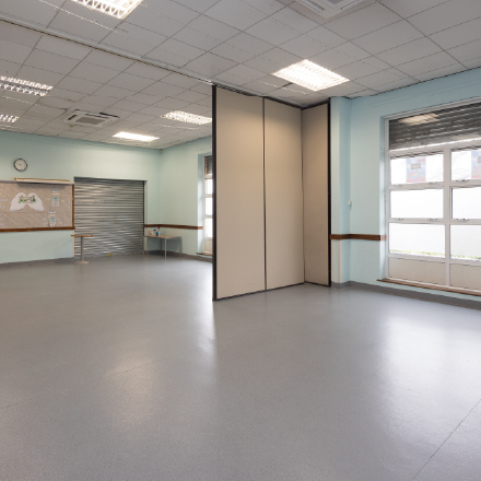 Spacious community hall with open floor area, movable partition wall, noticeboard, and large windows, suitable for events, classes, and group activities.