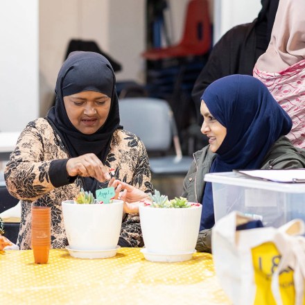 Two women in headscarves smile while arranging plants in white pots at a table covered with a yellow polka dot cloth.
