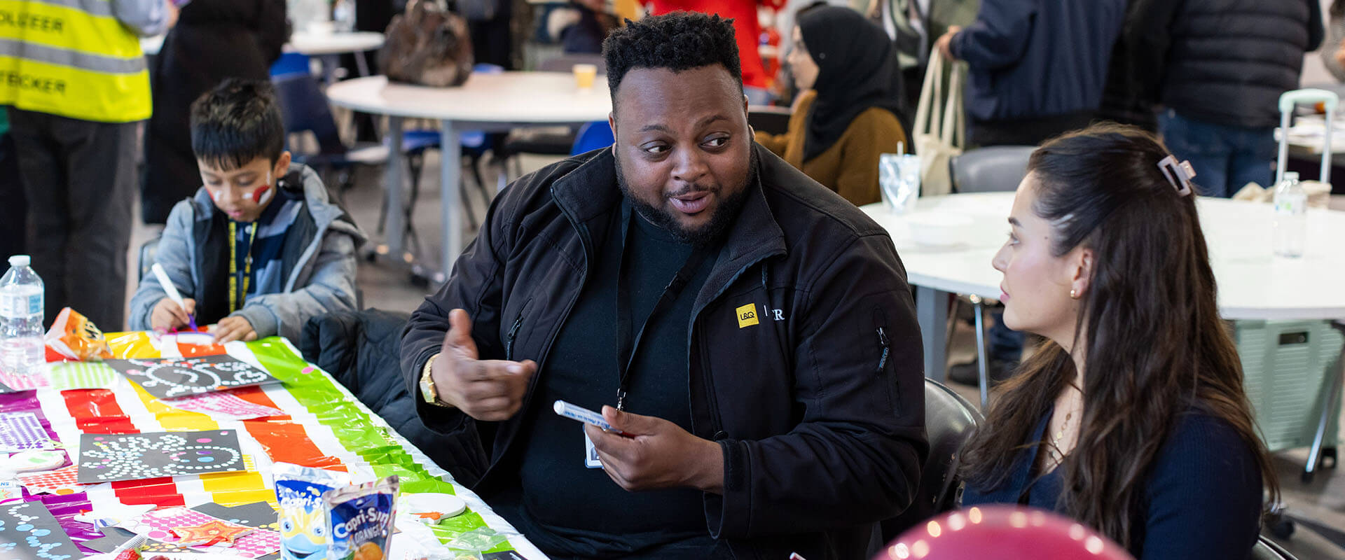 Man and woman talk at a colourful craft table while a child nearby works on an art project.