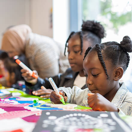 Young girl focuses on colouring while another child works beside her at a craft table.