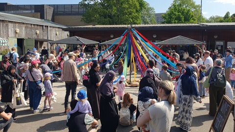 Large crowd of adults and children gathered outdoors around a colourful maypole decorated with long ribbons during a community celebration.