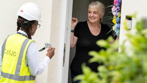 Cardo operative in high-visibility vest and hard hat approaching a resident in a black top standing at the front door.